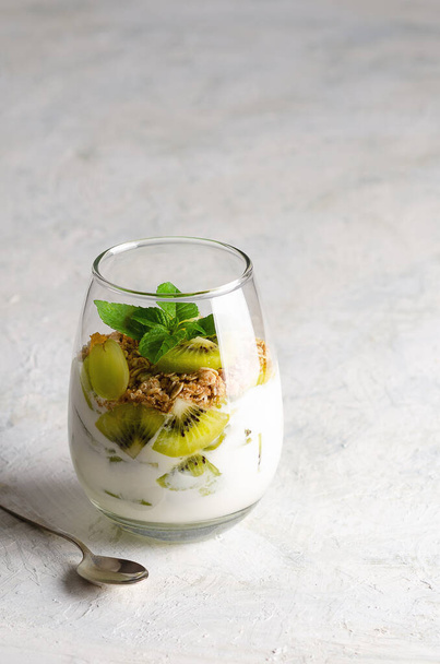 Plain yoghurt with pieces of fresh kiwi fruits, white grapes, granola, and mint leaves in a glass, on a grey background.  - Foto, Imagen