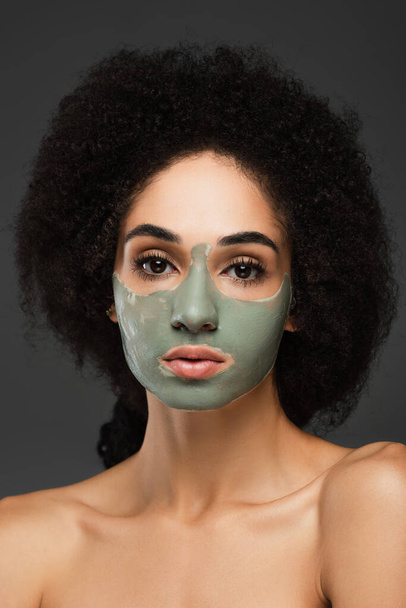 όμορφη Αφροαμερικανή γυναίκα με γυμνούς ώμους και πήλινη μάσκα προσώπου κοιτάζοντας την κάμερα που είναι απομονωμένη στο γκρι - Φωτογραφία, εικόνα