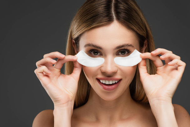 smiling woman looking at camera while holding eye patches isolated on grey - Foto, immagini
