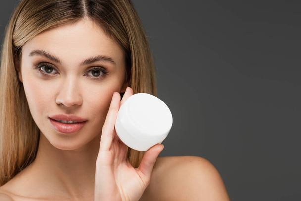 young woman with perfect skin and container of cosmetic cream looking at camera isolated on grey - Foto, Imagem