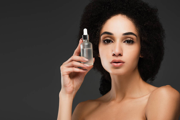 bella donna afroamericana con spalle nude in possesso di bottiglia di siero cosmetico isolato su grigio - Foto, immagini