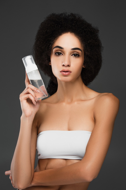 pretty african american woman in white top looking at camera while holding makeup remover isolated on grey - Photo, Image