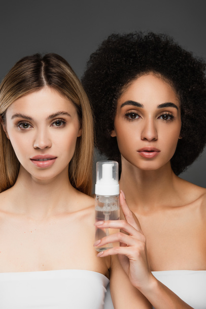 mujeres jóvenes y bastante multiculturales con la piel limpia mirando a la cámara cerca de espuma cosmética aislada en gris - Foto, imagen