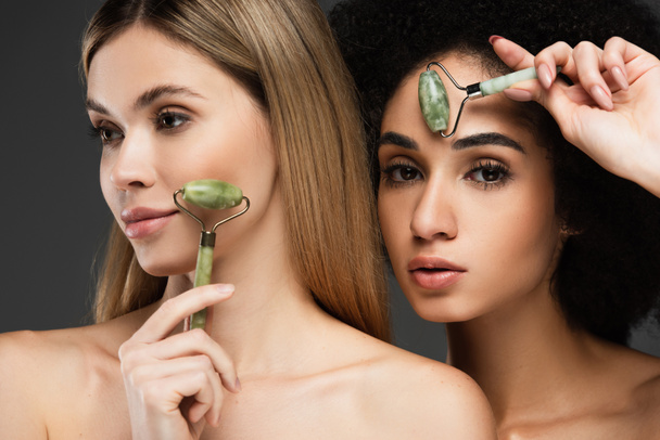 jovem interracial mulheres massagear rostos com jade rolos isolado no cinza - Foto, Imagem