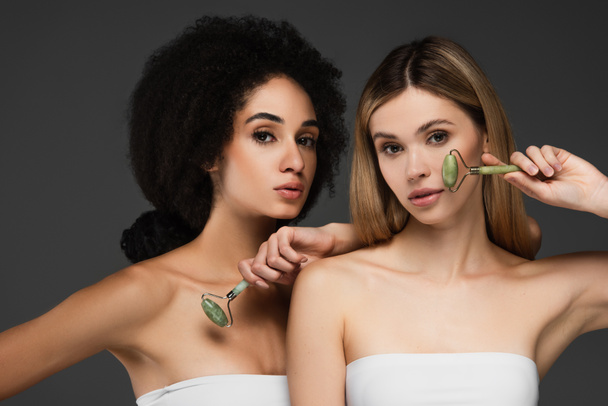 sensual interracial women with perfect skin and natural makeup posing with jade rollers isolated on grey - Photo, Image