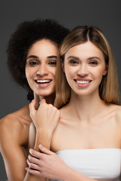 young and happy multiethnic women with naked shoulders smiling at camera isolated on grey - Zdjęcie, obraz