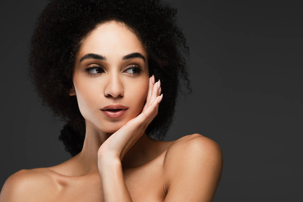 pretty african american woman with bare shoulder looking away while touching clean face isolated on grey - Photo, Image