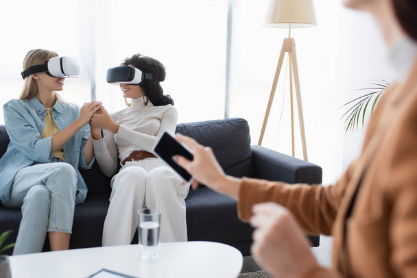 smiling same sex couple in vr headsets holding hands on sofa near blurred psychologist with smartphone  - Foto, Bild