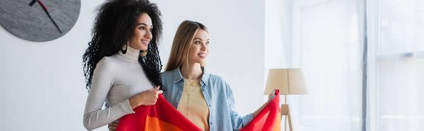 smiling interracial lesbian women looking away while holding lgbt flag, banner - Foto, afbeelding