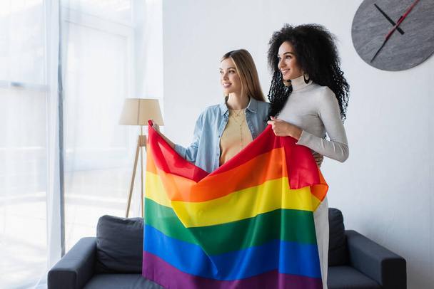 joyful interracial lesbian couple standing with lgbt flag near couch at home - Photo, Image