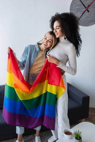 multiethnic lesbian girlfriends smiling while standing with lgbt flag - Foto, imagen