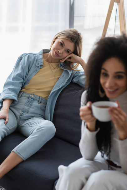 happy lesbian woman looking at blurred african american girlfriend drinking tea on couch - Foto, immagini