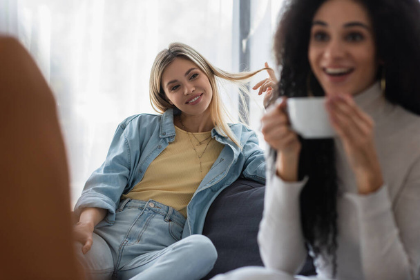 happy lesbian woman looking at blurred african american girlfriend drinking tea during psychological consultation - Foto, Bild