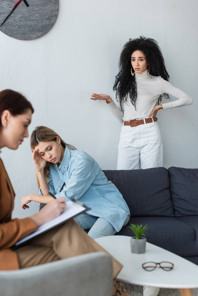 blurred psychologist writing on clipboard near depressed lesbian woman and her girlfriend standing with hand on hip - Foto, imagen