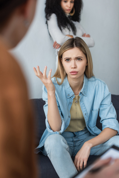 расстроенная лесбиянка разговаривает с размытым психологом рядом с африканской американской подружкой, стоящей со скрещенными руками - Фото, изображение