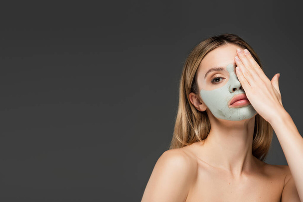 νεαρή γυναίκα με γυμνούς ώμους και πήλινη μάσκα σκιάζει το πρόσωπο με το χέρι απομονώνονται σε γκρι - Φωτογραφία, εικόνα