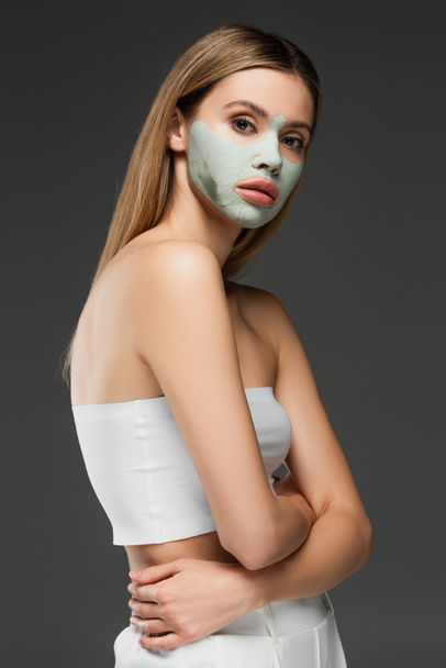 jong en slank vrouw verwennerij gezicht met klei masker geïsoleerd op grijs - Foto, afbeelding