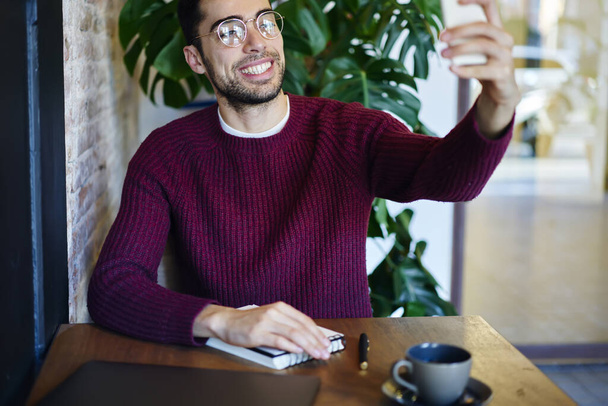 Cheerful bearded ethnic male in casual sweater and eyeglasses sitting at wooden table with cup of coffee and taking selfie - Valokuva, kuva