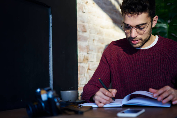 Concentrated young bearded male wearing warm knitted sweater and eyeglasses sitting at desk and writing notes in copybook while having coffee break - Fotografie, Obrázek