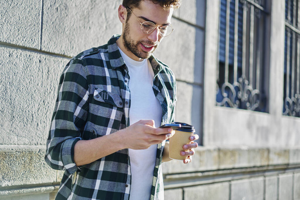 Young bearded male in casual clothes and eyeglasses browsing mobile phone during break from work and drinking coffee on street near modern building - Fotó, kép