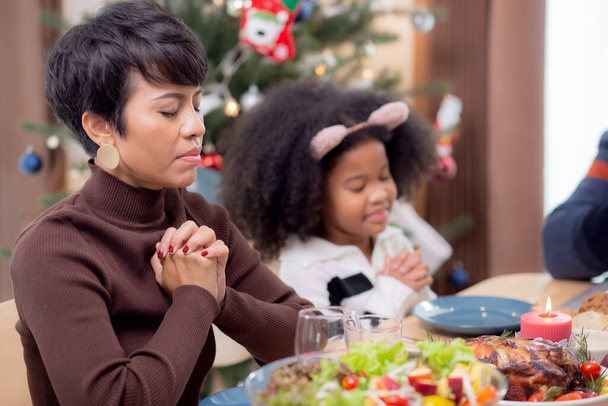 幸せなアフリカの家族が手を取り合って感謝祭の日に夕食を祈る一緒に、陽気なクリスマス、フードテーブルでのお祝いとお祝い、家族の関係、屋内、新年とクリスマス. - 写真・画像