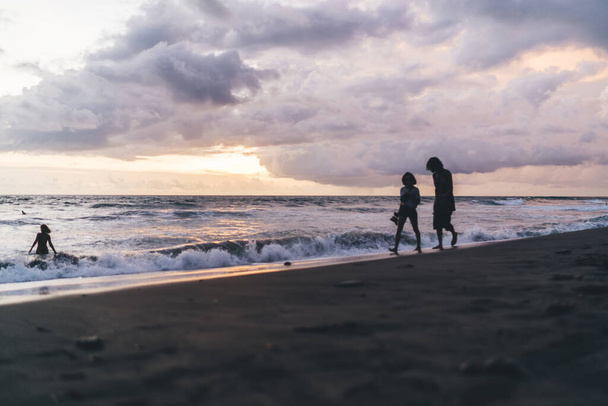 Full body barefoot unrecognizable silhouettes of people walking on sandy beach near waving sea during amazing sunset on Bali seashore - 写真・画像