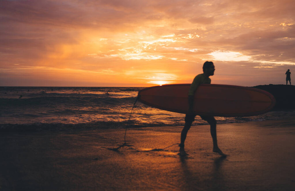 Side view of anonymous surfer with orange surfboard walking on empty sandy beach near water in evening during picturesque sunset - Photo, image