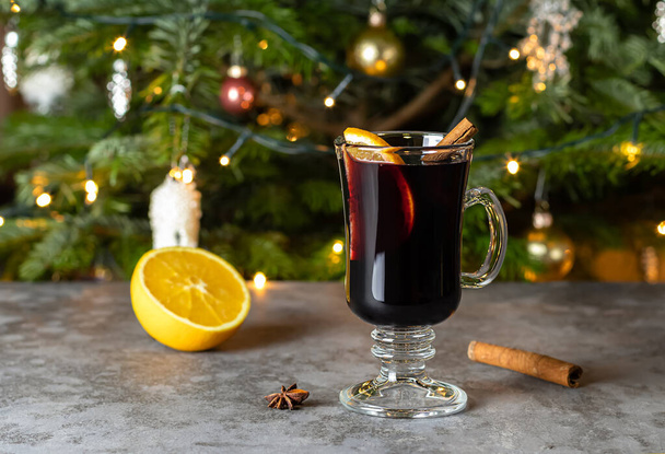 Sklenice vánočního svařeného vína nebo gluhweinu s kořením a pomerančovými plátky na stole proti rozmazanému vánočnímu stromečku.  - Fotografie, Obrázek