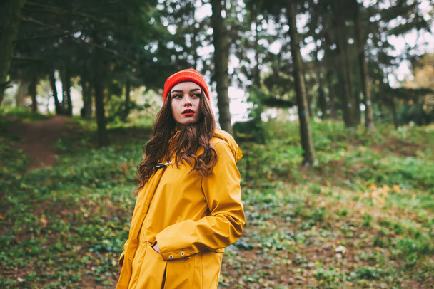 Young charming female in yellow raincoat and red hat with long wavy hair looking away while standing in forest with hands in pockets - Φωτογραφία, εικόνα