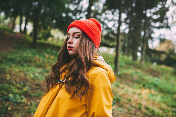 Side view of attractive female in yellow raincoat and hat thoughtfully looking down while standing on blurred background of green forest - Foto, Imagem