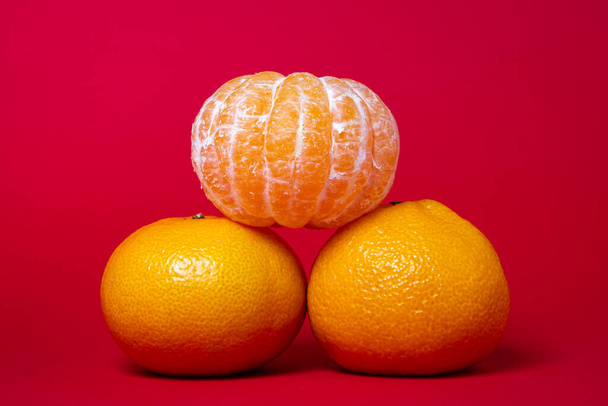 Peeled tangerine next to unpeeled tangerines on a red background. Citrus fruit on colored background - Foto, Imagem