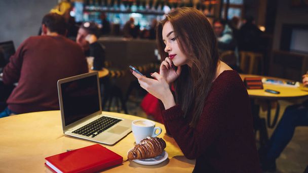 Side view of concentrated female sitting at table with laptop and cup of coffee while resting and surfing mobile phone - Photo, Image