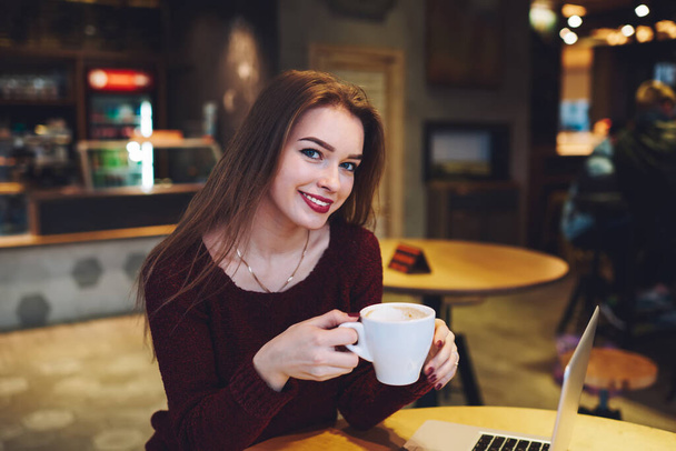 Positive female freelancer in casual clothes with toothy smile and red lips looking at camera sitting at table with laptop and cup of coffee - Foto, Imagen