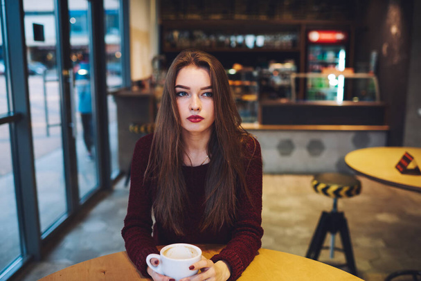 Focused female in casual clothes looking away sitting at round table with cup of coffee while having rest in cafe - Фото, изображение