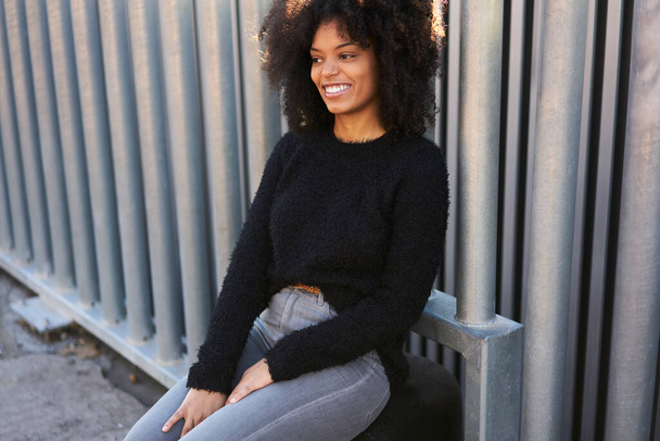 Crop young cheerful woman with curly hair in sweater smiling while sitting near metal fence on street in sunny day - Foto, afbeelding