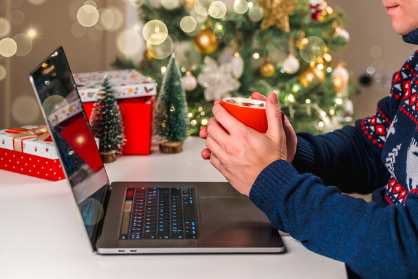 Ένας άντρας με γιορτινή μπλούζα πίνει κακάο με ζαχαρωτά κατά τη διάρκεια των γιορτών της Πρωτοχρονιάς και των Χριστουγέννων. Όμορφη ατμόσφαιρα των διακοπών στο σπίτι και στο χώρο εργασίας - Φωτογραφία, εικόνα