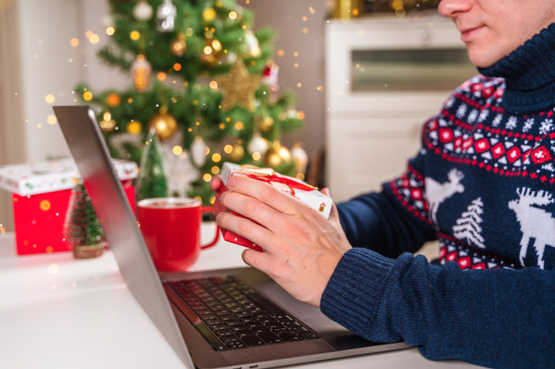 Een jongeman gekleed in een trui met een laptop aan zijn bureau naast een kerstboom en een gezellige nieuwjaarssfeer. Het concept van online winkelen en communicatie - Foto, afbeelding