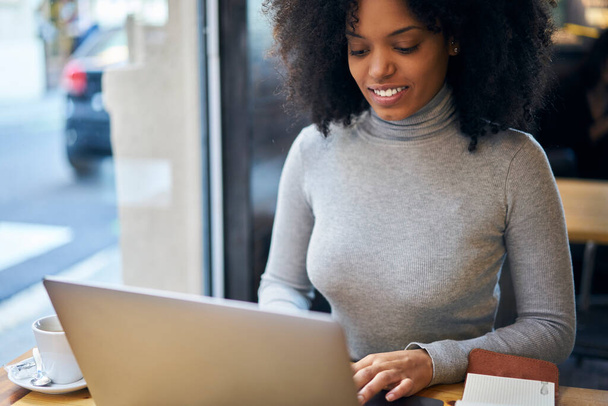 Crop young smiling African American female freelancer in casual outfit sitting at table in cafe and browsing netbook while working on project remotely - Foto, Imagem