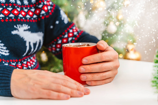 Ένας άντρας με γιορτινή μπλούζα πίνει κακάο με ζαχαρωτά κατά τη διάρκεια των γιορτών της Πρωτοχρονιάς και των Χριστουγέννων. Όμορφη ατμόσφαιρα των διακοπών στο σπίτι και στο χώρο εργασίας - Φωτογραφία, εικόνα