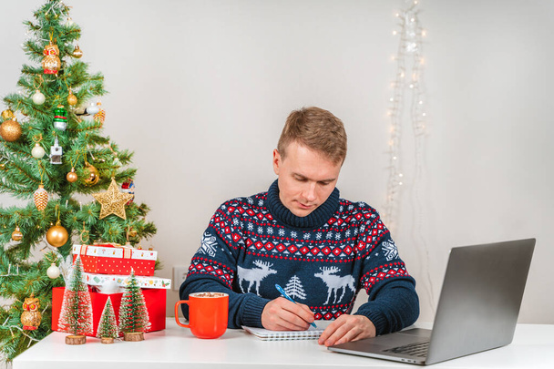 Młody mężczyzna ubrany w sweter, korzystający z laptopa przy biurku obok choinki i przytulnej atmosfery Nowego Roku. Koncepcja zakupów i komunikacji internetowej - Zdjęcie, obraz