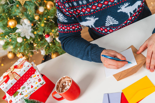  Un joven con un suéter firma postales y escribe cartas junto a un árbol de Navidad. El concepto de preparación para la celebración de Año Nuevo y Navidad - Foto, imagen