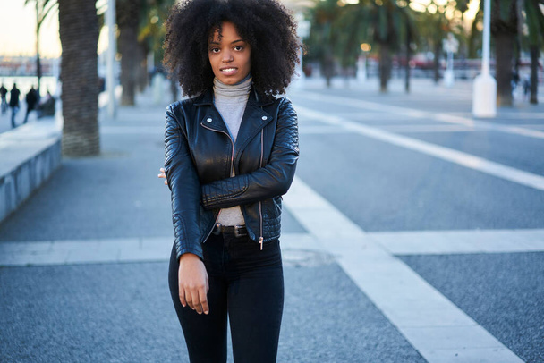 Serious African American female in stylish outfit standing on street and looking at camera while resting after walking in city - Photo, Image