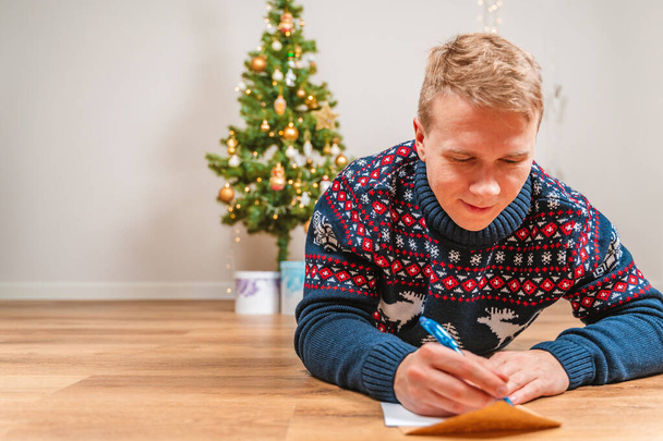  Un joven en un suéter yace en el suelo frente a un árbol de Navidad y escribe una carta - Foto, imagen