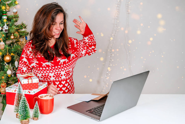 Nuori nainen punaisessa puserossa käyttää kotona kannettavaa tietokonetta kommunikoidakseen videoneuvottelun kautta joululomien aikana. Tyttö omistaa joululahjan ja kiittää ystäviä tai sukulaisia - Valokuva, kuva