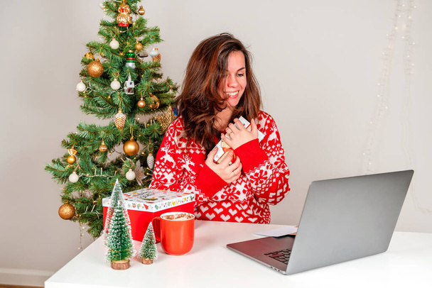 Eine junge Frau im roten Pullover nutzt zu Hause einen Laptop, um über die Weihnachtsfeiertage per Videokonferenz zu kommunizieren. Ein Mädchen hält ein Weihnachtsgeschenk in der Hand und dankt Freunden oder Verwandten - Foto, Bild