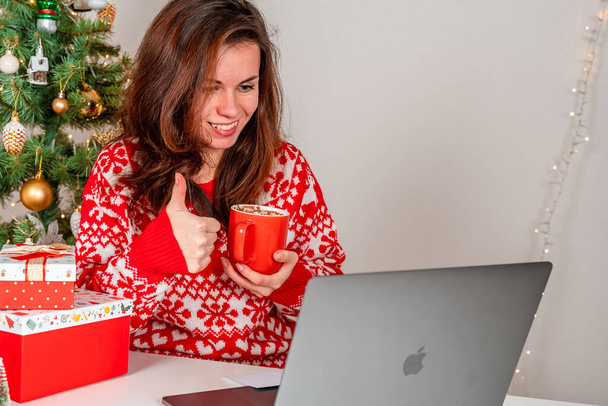 Μια νεαρή γυναίκα με κόκκινο πουλόβερ χρησιμοποιεί ένα φορητό υπολογιστή στο σπίτι για να επικοινωνεί μέσω τηλεδιάσκεψης κατά τη διάρκεια των εορτών των Χριστουγέννων. Ένα κορίτσι κατέχει ένα χριστουγεννιάτικο δώρο και ευχαριστεί τους φίλους ή τους συγγενείς - Φωτογραφία, εικόνα