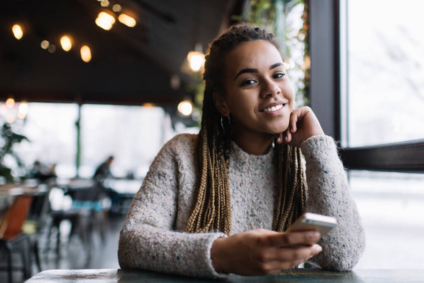 Portrait of cheerful hipster girl with mobility technology in hand resting at cafeteria table, happy millennial Generation Z with Afro hairstyle smiling at camera during smartphone networking - Photo, image
