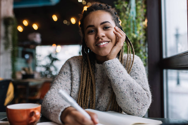 Portrait of pretty hipster girl with cute smile on face posing at cafeteria table while working with poetry ideas, carefree woman with pigtails braiding enjoying studying time for doing homework - Foto, Bild