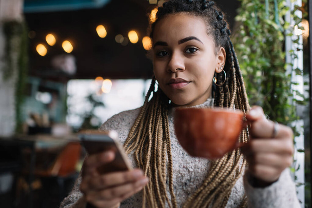 Portrait of African American female customer sitting at cafe table with aroma cappuccino holding modern smartphone and looking at camera, beautiful millennial woman with cellular device posing - Photo, image