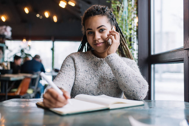Pretty African American student enjoying smartphone talking during learning education in coffee shop, portrait of intelligent female blogger with planning textbook using cellular for online phoning - Foto, immagini